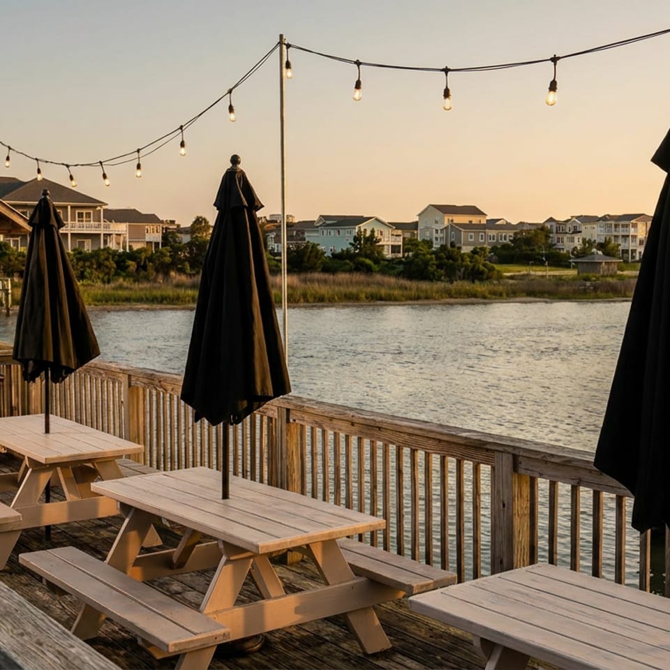 Waterfront restaurant deck with wooden picnic tables, closed black umbrellas, string lights, boats, and coastal homes at sunset.
