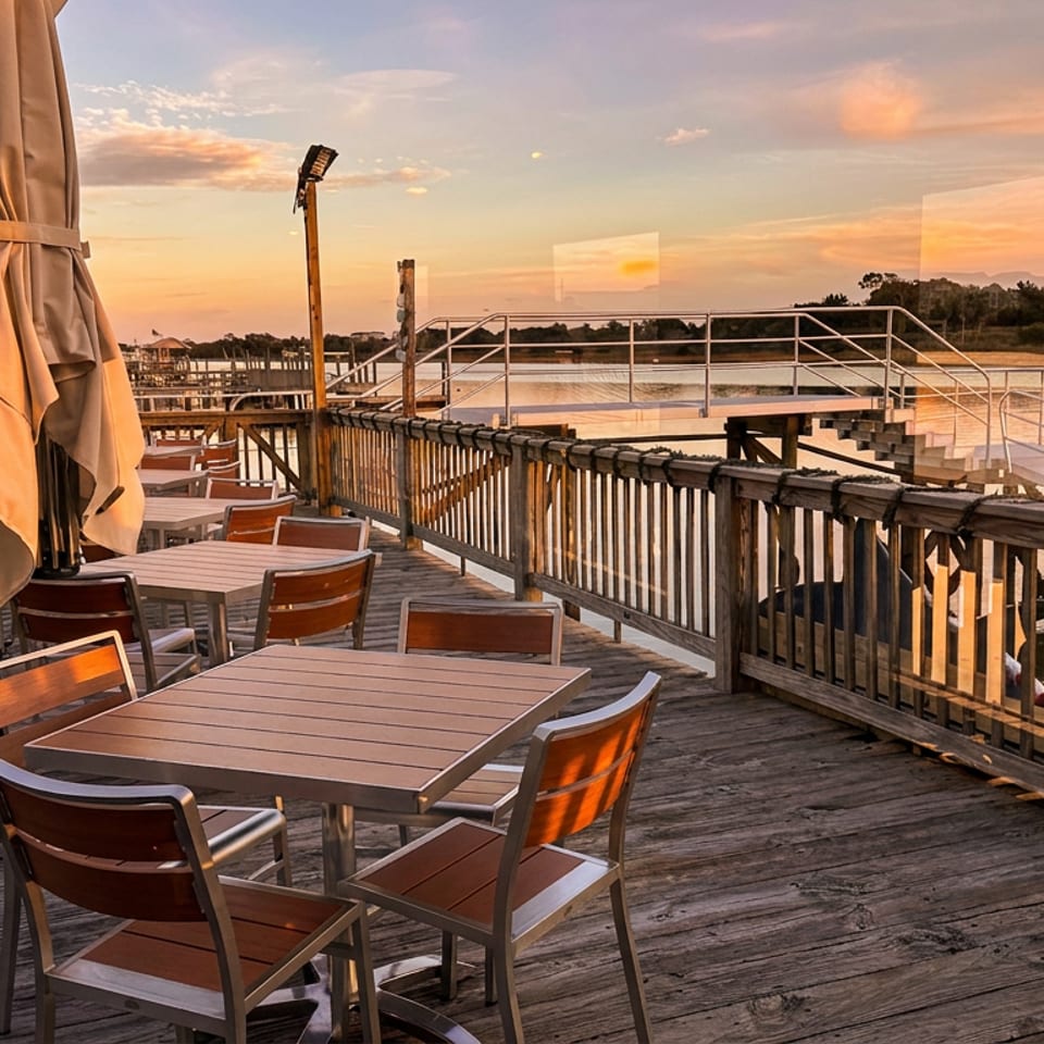 Waterfront restaurant deck with outdoor tables, chairs, umbrellas, and a dock overlooking calm waters at sunset.