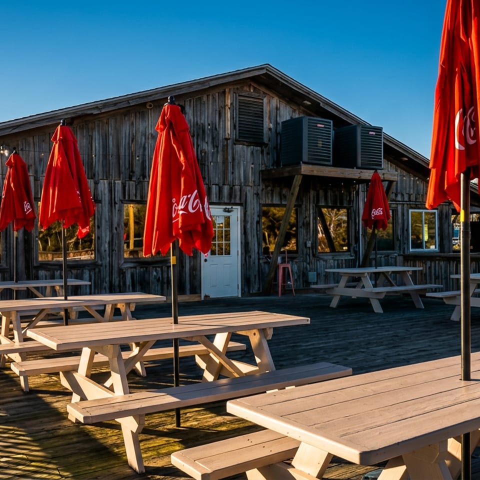 Outdoor waterfront restaurant deck with wooden picnic tables, closed red Coca-Cola umbrellas, string lights, and boats visible beyond.