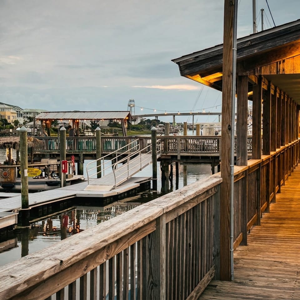 Covered wooden waterfront restaurant walkway overlooks docks, boats, and calm water beneath a cloudy evening sky.