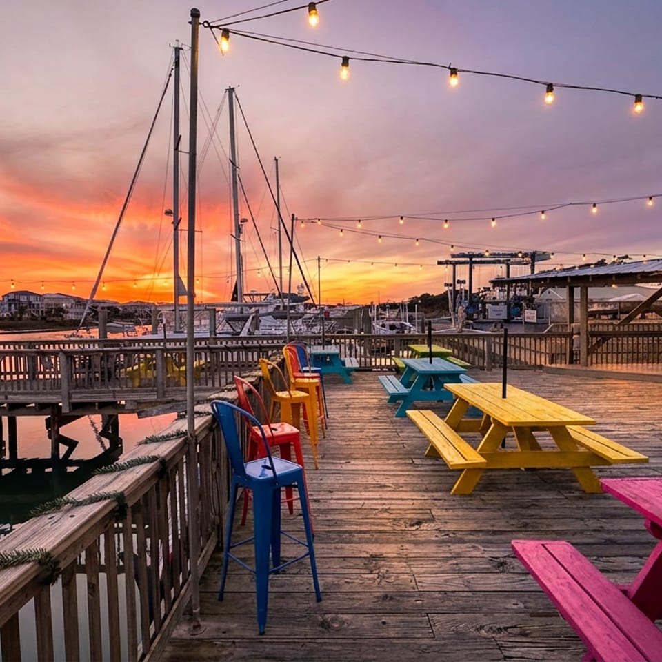 Colorful picnic tables and bar chairs on a waterfront restaurant deck beneath string lights at sunset, with boats and docks beyond.