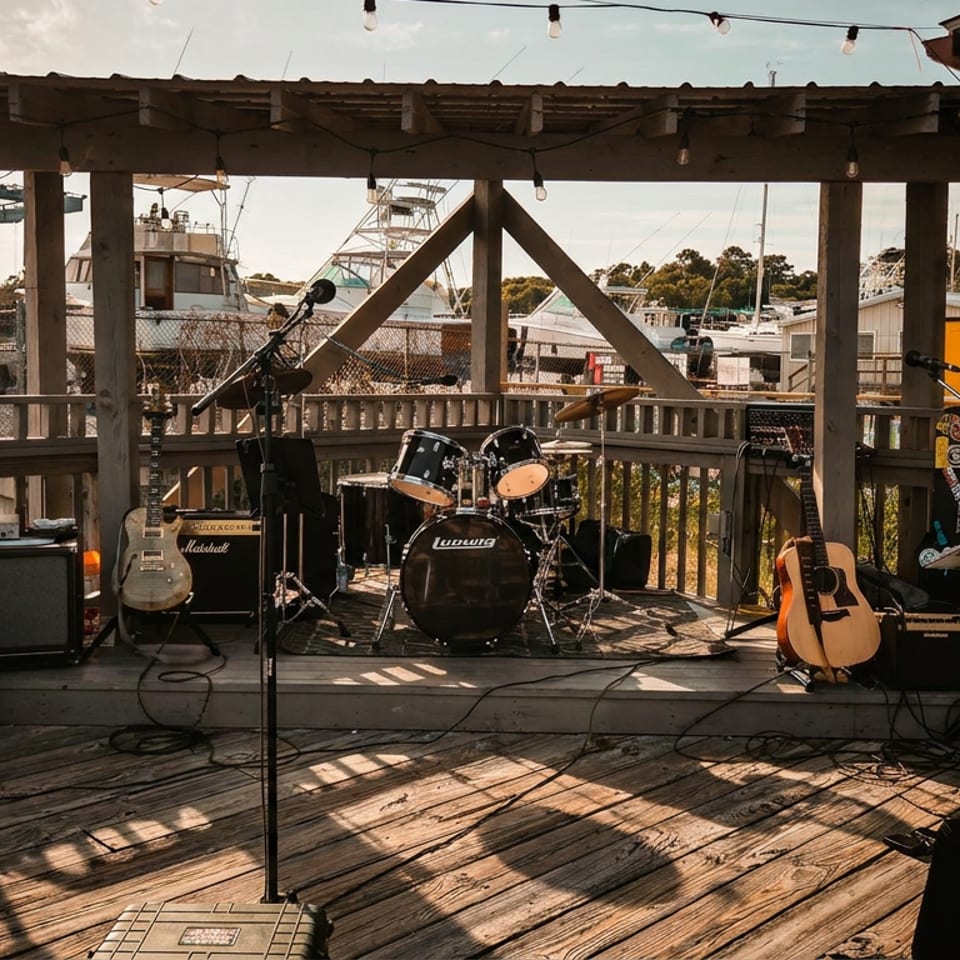 Outdoor waterfront restaurant stage with guitars, drum kit, microphones, speakers, string lights, and fishing boats in the harbor beyond.
