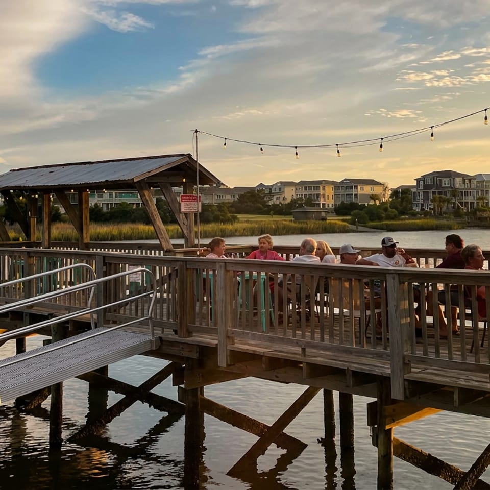 Diners gather on a waterfront restaurant deck overlooking calm waters and seaside homes at sunset, with string lights overhead.