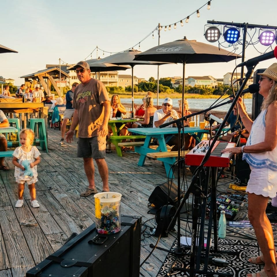 Live music performance on a waterfront restaurant deck, with a keyboardist, dancing guests, picnic tables, umbrellas, and string lights.
